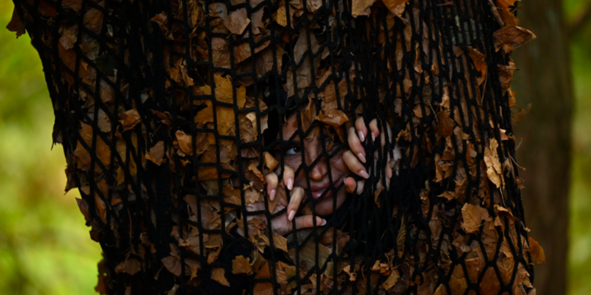 Photo of Mercedes "MJ" Javid peeking through a leaves while she is held in a net from episode seven of season two of The Traitors