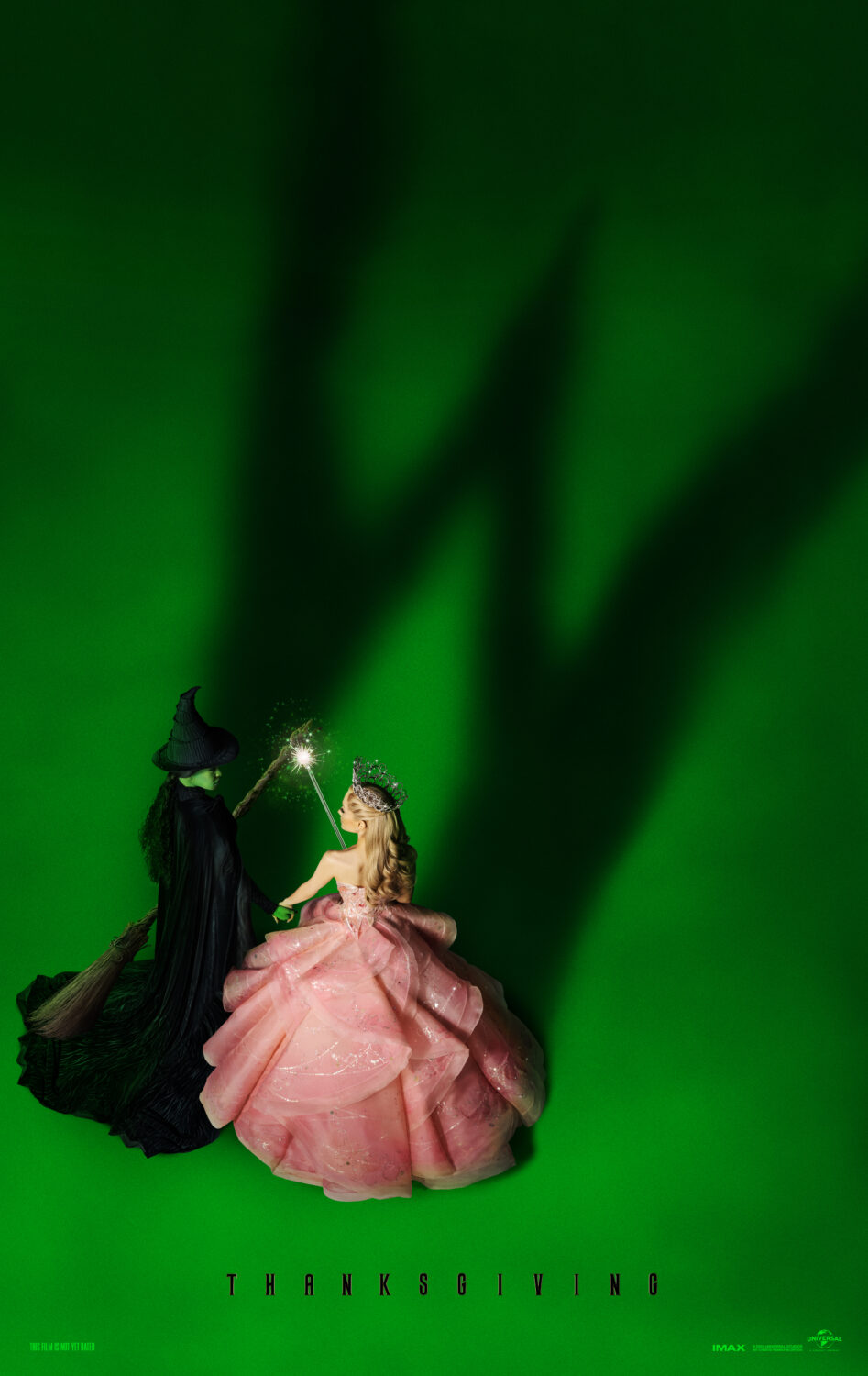 Poster for Wicked the movie, green background with small figurines of Elphaba and Glinda with their shadow making a W
